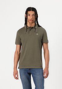 Olivgrön polo t-shirt i bomull med krage och två knappar, med en liten broderad logotyp på bröstet. Bärs med blå jeans.