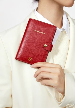 Persona con blazer blanco sostiene una cartera texturizada roja de Victoria Hyde con botón de presión y letras doradas que dicen "Ama lo que haces."