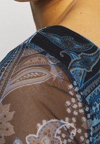 Top transparente con un patrón de cachemira en tonos azules y marrones, con tela negra sólida a lo largo de las costuras de los hombros y el escote.