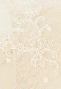 Tessuto di pizzo beige delicato con ricami floreali e piccoli punti di texture. Una cucitura bianca intricata forma un motivo dettagliato.