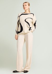 Maglione beige con audaci linee astratte nere, abbinato a pantaloni crema a gamba larga e mocassini neri lucidi, che mostrano una vestibilità rilassata.