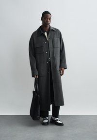 Garšs pelēks vilnas mētelis ar plašu apkakli, četriem priekšējiem kabatām un spiedpogu aizdari. Valkāts virs melna biksēm un baltām kurpēm. Turēja melnu rokassomu.