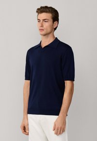 Joven con una camisa tipo polo de manga corta azul marino y pantalones blancos, de pie frente a un fondo liso de color gris claro.