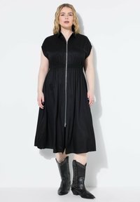 Abito nero con zip, maniche corte, vita arricciata e gonna fluida. Un paio di stivali cowboy neri con borchie aggiunge contrasto.