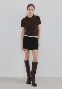 Mujer con suéter de manga corta marrón oscuro, minifalda asimétrica negra y botas marrón oscuro hasta la rodilla, de pie contra una pared lisa.