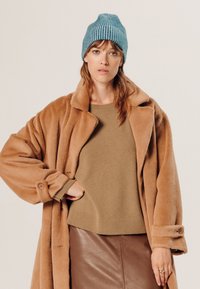 Bruine nepbontjas, beige gebreide trui en een bruine leren rok. Blauwe geribde beanie op het model; zachte texturen en gelaagd ontwerp.