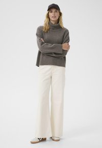 Brown turtleneck sweater with ribbed cuffs, paired with wide-legged cream pants and white sneakers. The model wears a black cap.