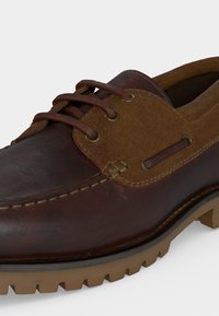 Καφέ δερμάτινο και σουέτ boat shoe με καφέ κορδόνια και ανθεκτική ταμπά λαστιχένια σόλα, υπό γωνία που δείχνει τις ραφές και τα οπές για τα κορδόνια.