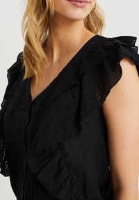 Musta paita, joka on valmistettu kevyestä kankaasta ja jossa on röyhelösateet, V-mallinen kaula-aukko, pleated-pinta ja napitettu etuosa.