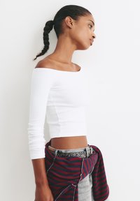 Bílý žebrovaný dlouhý rukáv crop top, doplněný červeným a námořnickým pruhovaným svetr, který je uvázaný kolem pasu, šedé džíny a opasek se cvočky.