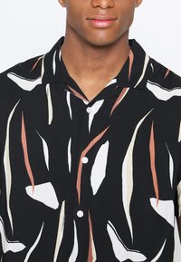 Chemise noire à boutons présentant un motif de formes abstraites en blanc crème, rouille et beige, avec un col décontracté et une texture lisse.