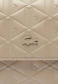 Portefeuille en cuir beige texturé présentant un motif géométrique en relief avec un petit logo de crocodile Lacoste en relief.
