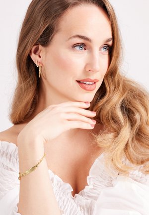 Des boucles d'oreilles créoles en or et un bracelet en chaîne doré sont portés ; le modèle a de longs cheveux ondulés et clairs et est vêtu d'un haut blanc à volants.