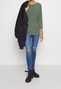 Grünes Langarmoberteil, blaue zerrissene Skinny Jeans und schwarze Bomberjacke. Klobige schwarze Knöchelstiefel mit Schnallen. Glatter Stoff und lockere Passform.