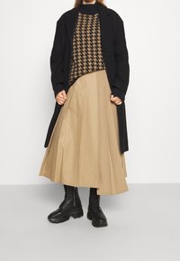 Persoon draagt een zwarte lange jas, een bruin-zwart pied-de-poule trui, een beige geplooide midi-rok en zwarte vetercombatlaarzen.