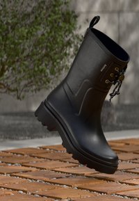 Schwarzer Regenstiefel bis zum Knöchel mit dicker Profilsohle und Schnürdetails, schwebend über braunen Holzklötzen mit kleinen grauen Steinen.