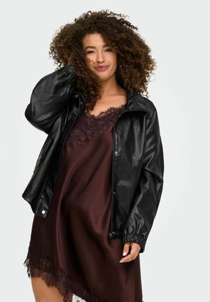 Une femme aux cheveux bouclés pose devant un fond uni, vêtue d’une veste en cuir noire par-dessus une robe bordeaux bordée de dentelle.