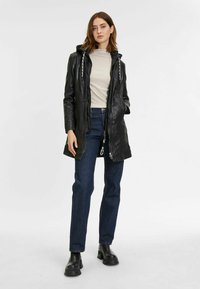 Cappotto lungo in pelle nera con cappuccio, dettagli in metallo argentato, due tasche frontali e chiusura con cordino. Indossato sopra un top chiaro e jeans.