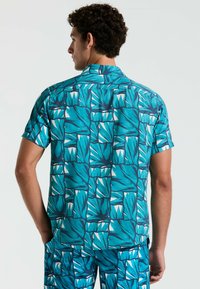 Korte mouwen shirt in turquoise met een levendig bladdessin, voorzien van een kraag en detail op de rug. Geweven stof voegt diepte toe.