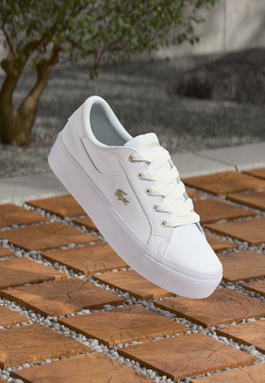 White synthetic sneakers with lace-up design, featuring a textured white sole and a gold logo accent. Set against a brown tiled background.