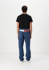 T-shirt noir, pantalon cargo en denim bleu avec de nombreuses poches et une ceinture contrastante, ornés d'accents rouges et bleus. Baskets blanches.