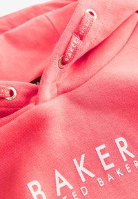 Sudadera color coral con una textura de tela suave, ojales de metal y letras impresas en blanco en la parte delantera. Los cordones también son color coral.