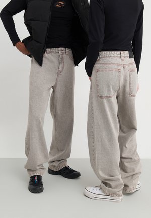 Zwei Personen tragen hellgraue Jeans mit weitem Bein und kontrastierenden roten Nähten, schwarze und weiße Sneaker und schwarze Oberteile.