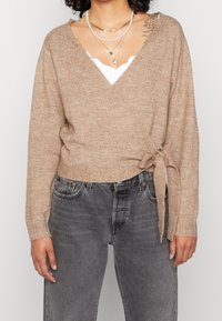 Cardigan avvolgente beige a maglia con maniche lunghe, dettaglio legato in vita, indossato sopra una maglietta bianca, abbinato a jeans scuri grigi a vita alta.