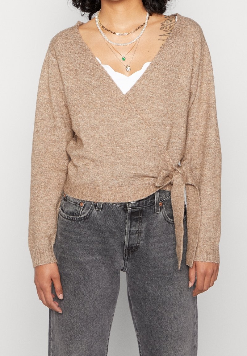 Cardigan avvolgente beige a maglia con maniche lunghe, dettaglio legato in vita, indossato sopra una maglietta bianca, abbinato a jeans scuri grigi a vita alta.