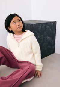 Sweat à capuche blanc côtelé avec fermeture éclair et capuche, porté par-dessus un t-shirt rose clair, associé à un pantalon de survêtement rose ample, assis sur une surface lisse.