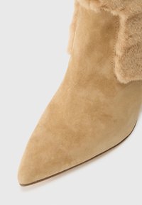 Botine din suede bej, cu vârful ascuțit, bordură moale din blană în jurul gleznei și detalii fine de cusătură.