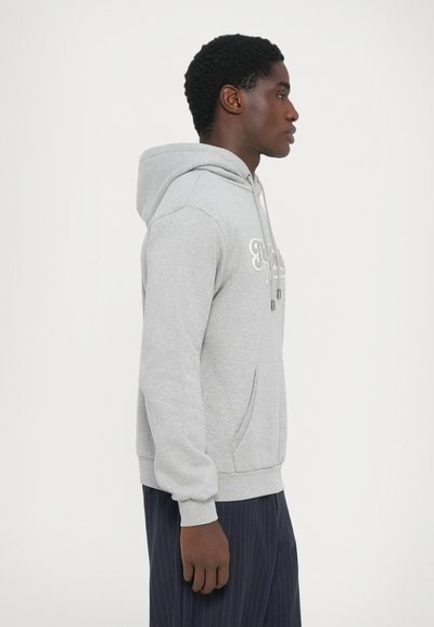 Un hoodie gris avec une poche avant et une capuche à cordon. Présente un texte brodé sur la poitrine. Porté avec un pantalon bleu marine à rayures.