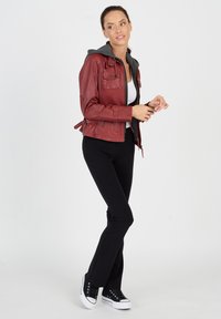 Rote Lederjacke mit grauem Kapuzenpulli, Reißverschluss und mehreren Taschen. Schwarze Hose und schwarze Sneakers runden das Outfit ab.