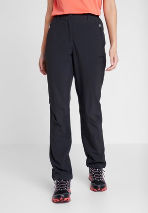 Osoba ubrana w czarne spodnie outdoorowe i czarno-czerwone buty turystyczne, stojąca na jednolitym jasnoszarym tle.