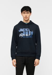 Νεαρός άνδρας φοράει μπλε σκούρο φούτερ με κουκούλα με το κείμενο "Jack & Jones" και άλλα κείμενα σε λευκό και μπλε, συνδυασμένο με μαύρο παντελόνι, στέκεται όρθιος.