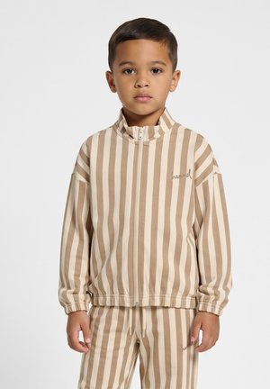 Jeune garçon portant une veste zippée beige et blanche à rayures verticales et un pantalon assorti, debout devant un fond clair uni.