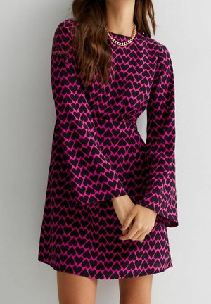 Femme portant une robe à manches longues violette avec un motif de cœurs noirs et un collier en chaîne dorée, les mains jointes devant elle.