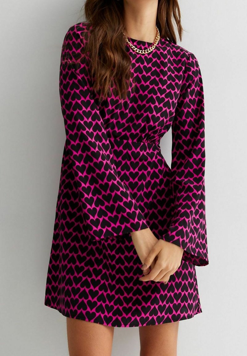 Femme portant une robe à manches longues violette avec un motif de cœurs noirs et un collier en chaîne dorée, les mains jointes devant elle.