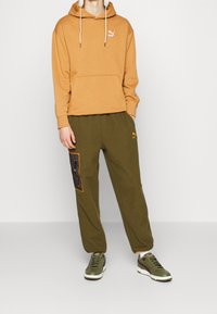 Felpa marrone con tasca a canguro e logo bianco, abbinata a pantaloni della tuta verde oliva con una toppa decorata e polsini elasticizzati. Sneakers verdi.