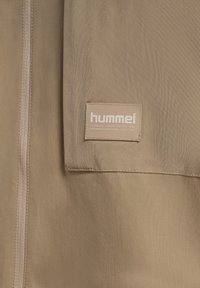 Beżowa tkanina w zbliżeniu, na której widać zamek błyskawiczny oraz prostokątną naszywkę z logo marki "hummel" i tekstem o odzieży technicznej i sportowej.