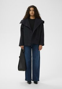 Schwarze, übergroße Wolljacke mit breiten Revers, doppelreihiger Vorderseite, kombiniert mit ausgestellten blauen Jeans und einer schwarzen Einkaufstasche.