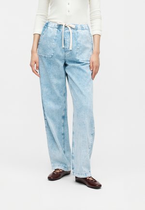 Γυναίκα που φοράει ανοιχτό μπλε τζιν παντελόνια με φαρδιά πόδια, κορδόνι στη μέση και μεγάλες μπροστινές τσέπες, σε συνδυασμό με λευκό τοπ και καφέ φλατ παπούτσια.