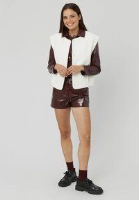 Gilet blanc duveteux sur une chemise en vinyle bordeaux et un short. Chaussures noires à lacets avec une semelle épaisse. Fond gris clair.