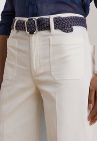 Pantalons blancs taille haute avec de grandes poches angulaires. Une ceinture tissée bleu foncé avec une boucle argentée brillante maintient la taille.