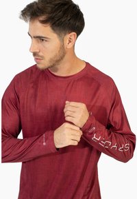 Pitkähihainen urheilupaita maroonissa, jossa on teksturoitu pinta, pyöreä kaula-aukko ja logo hihassa. Kangas näyttää kevyeltä ja hengittävältä.