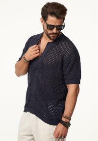 Marinefarbener Kurzarm-Cardigan, offenes Strickdesign mit großen Löchern, Knopfverschluss. Über hellen Hosen getragen, accessorized mit Armbändern.