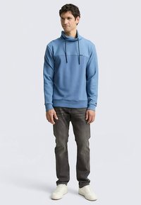 Sweat-shirt bleu clair avec un col montant et des cordons de serrage, présentant une couture horizontale sur la poitrine et des poignets côtelés, assorti à un jean gris.