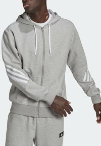 Felpa con cappuccio grigia con zip e coulisse, caratterizzata da dettagli a tre strisce bianche sulle maniche e una tasca a marsupio. Morbido tessuto misto cotone.