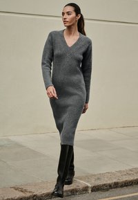 Robe grise en maille côtelée à manches longues avec décolleté en V, silhouette ajustée et fini texturé, associée à des bottes en cuir noir montant jusqu'au genou.