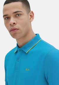 Polo shirt turchese realizzato in tessuto morbido, con colletto a coste e dettagli gialli, e un piccolo logo giallo sul petto.